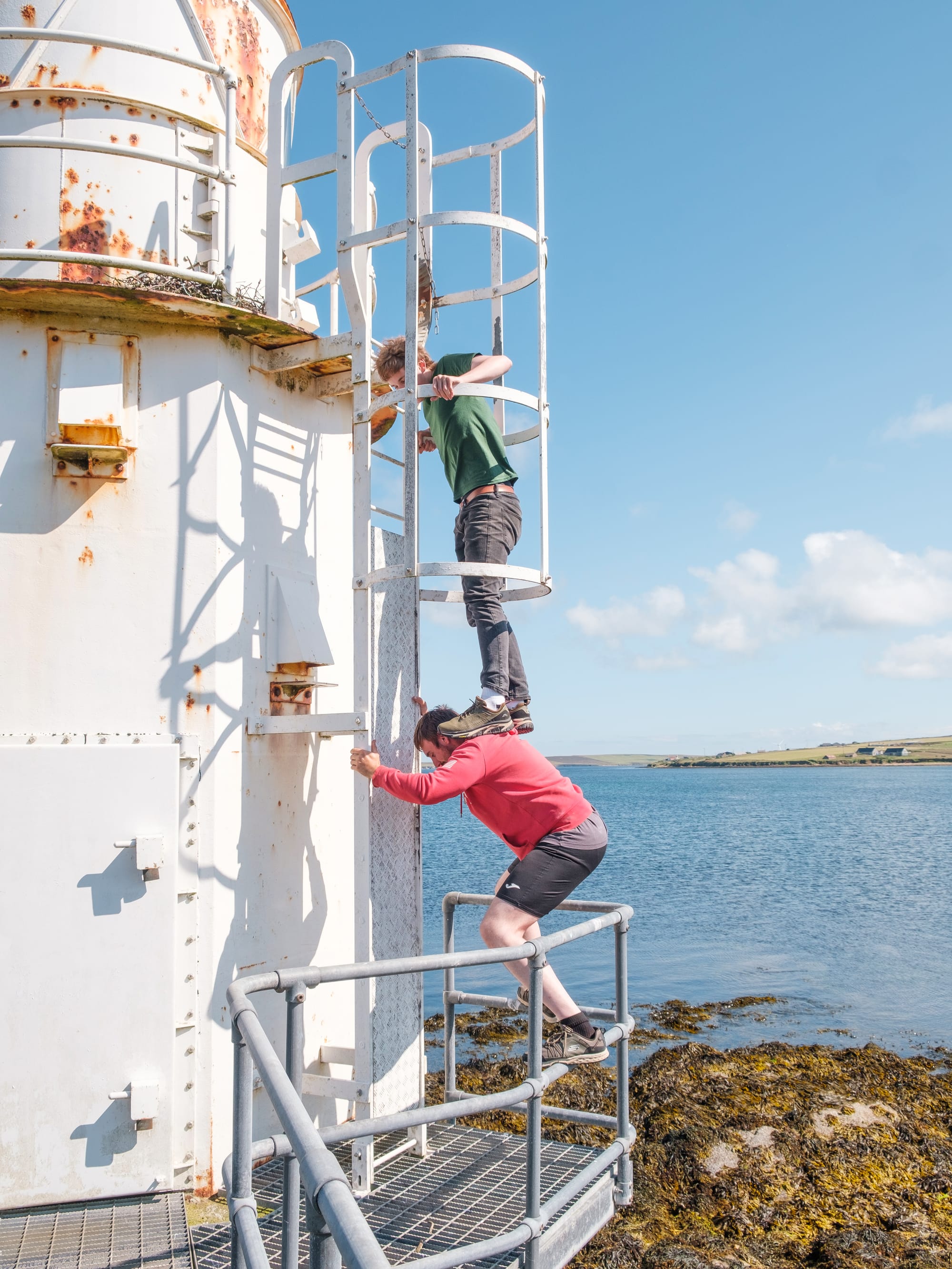 Two people climb a rusted lighthouse ladder above the rocky shore.Two people climb a rusted lighthouse ladder above the rocky shore.