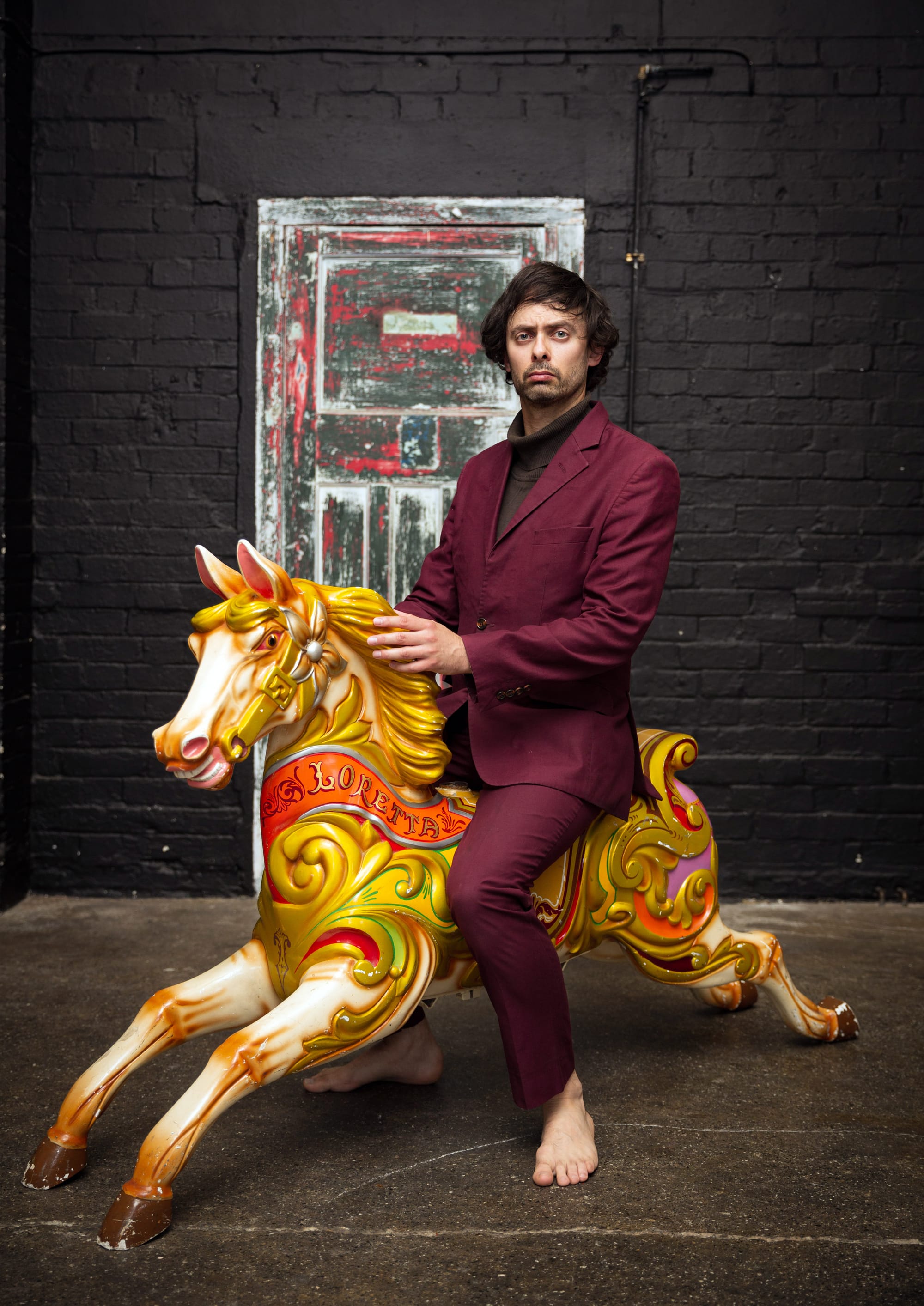 Marcel Lucont in a burgundy suit sits barefoot on a painted fairground horse indoors.