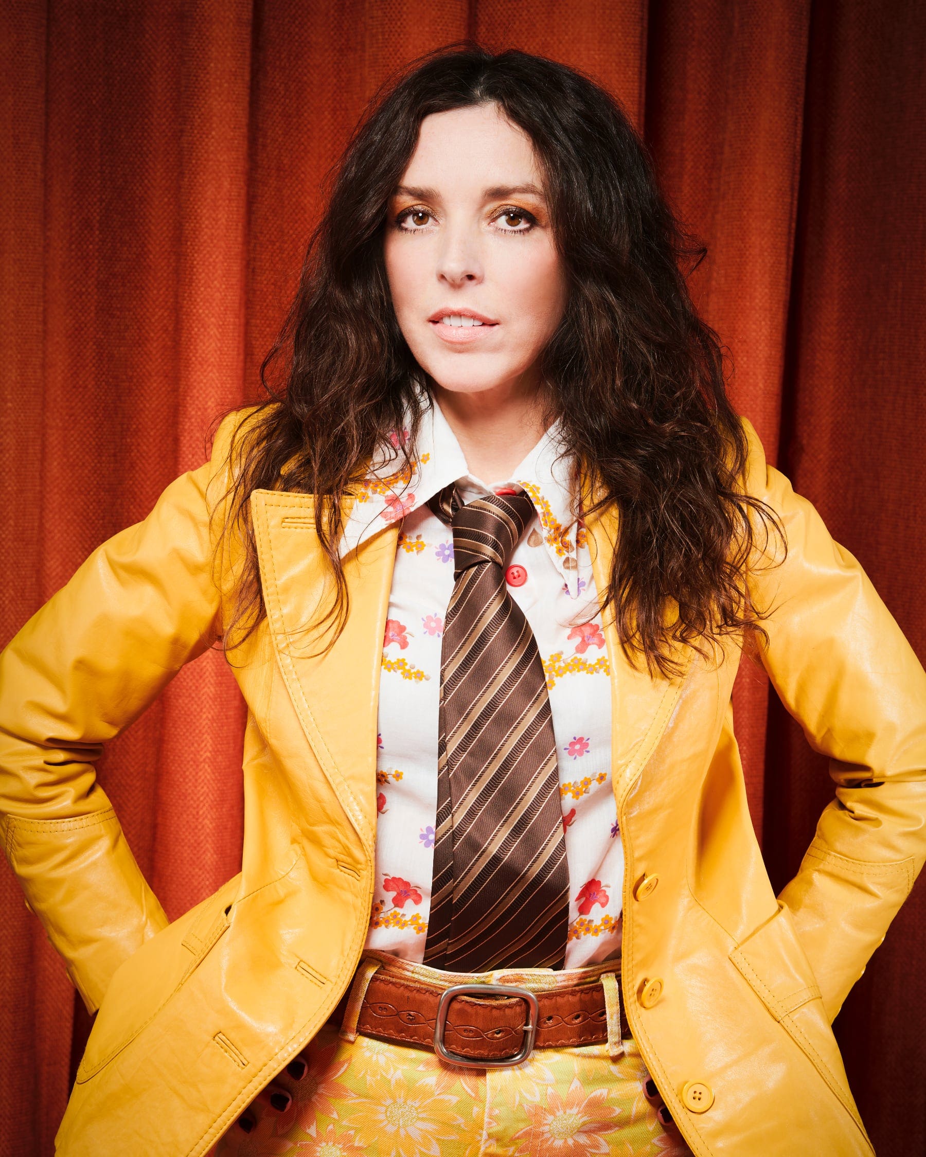 Bridget Christie wears a yellow suit and patterned tie, standing confidently in front of red curtains.