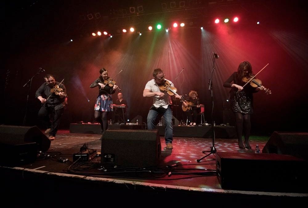 Five musicians play fiddles on stage under colourful lights, with piano and guitar behind them.