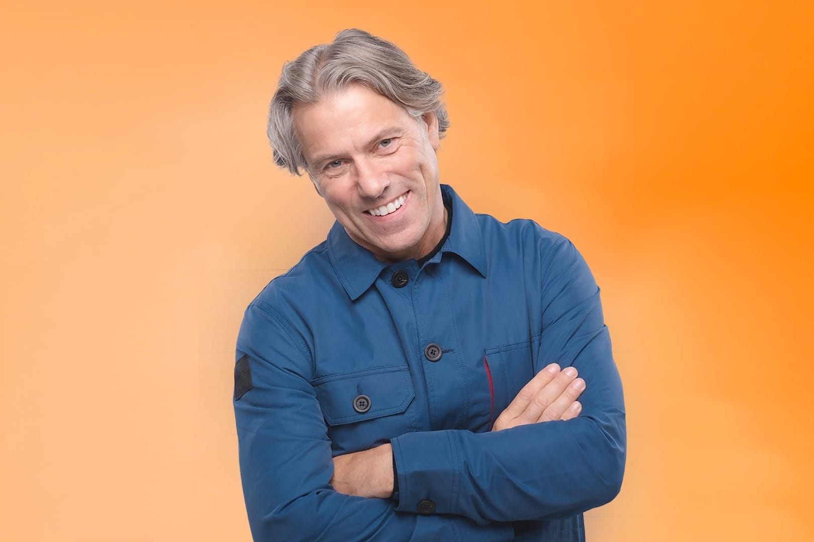 John Bishop smiles with arms crossed, wearing a blue jacket against an orange background.