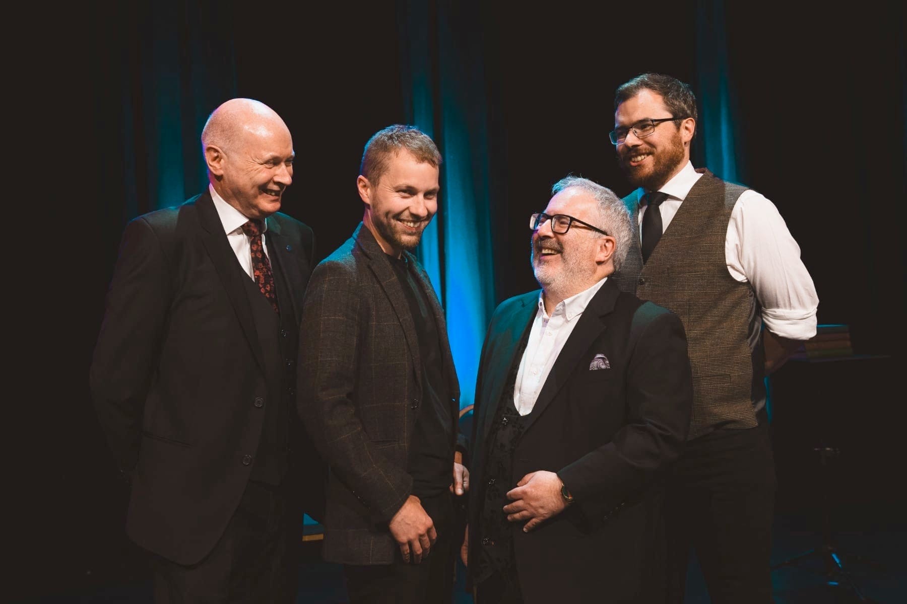 our men in suits and waistcoats stand laughing together on stage under warm lighting.
