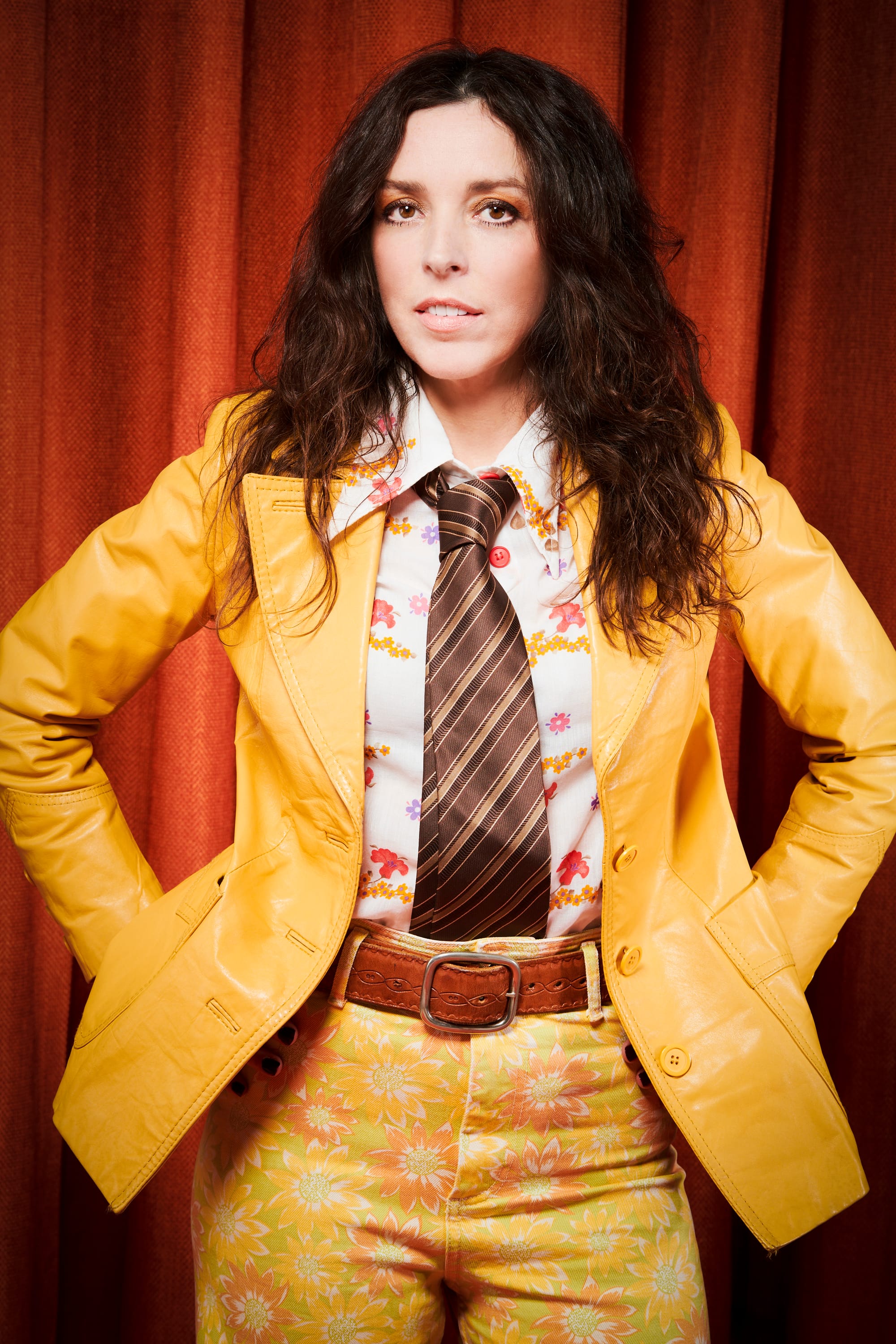 Bridget Christie stands in front of red curtains wearing a yellow jacket, floral trousers, and a striped tie.