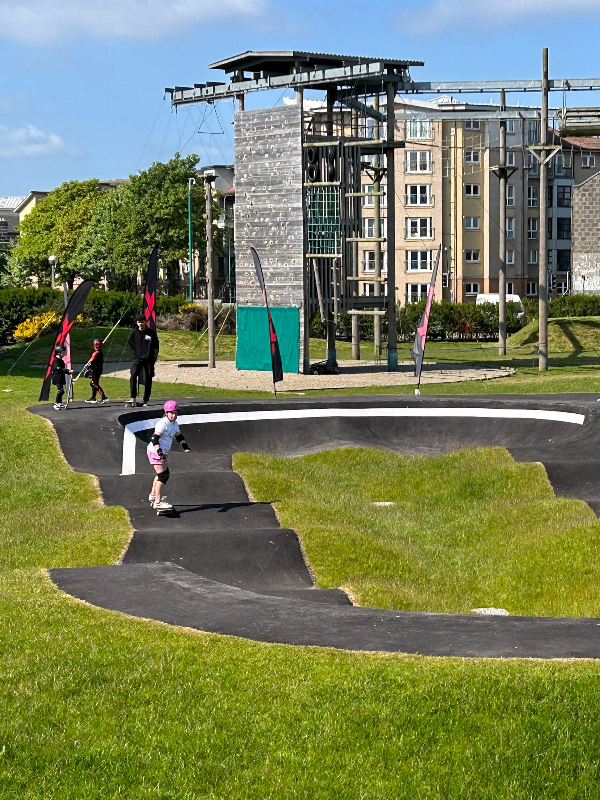 Child wearing a helmet skateboards on an outdoor pump track near climbing structures.