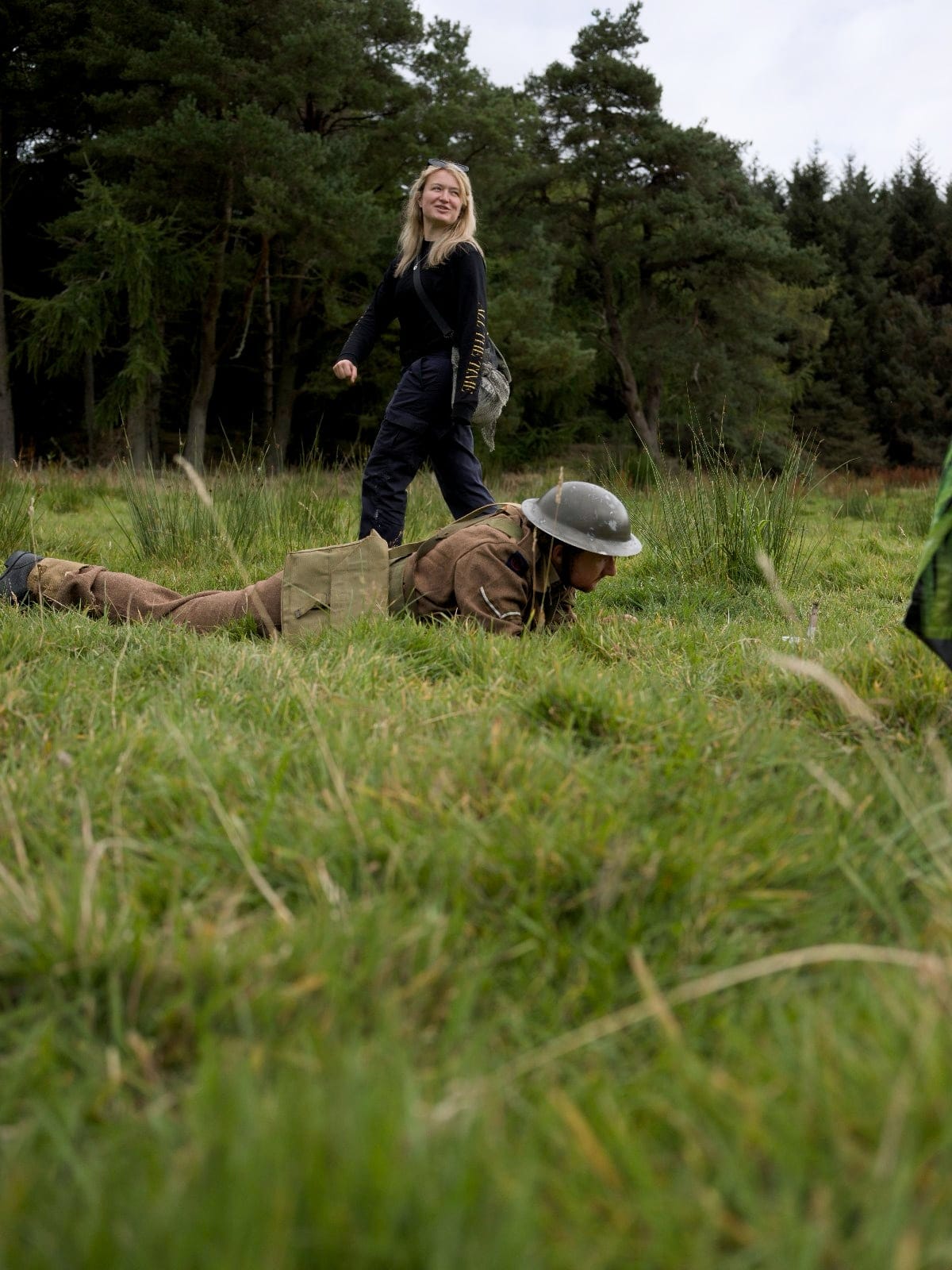 Phoebe Banks walks through grassy woodland while an actor in a soldier’s uniform crawls nearby.