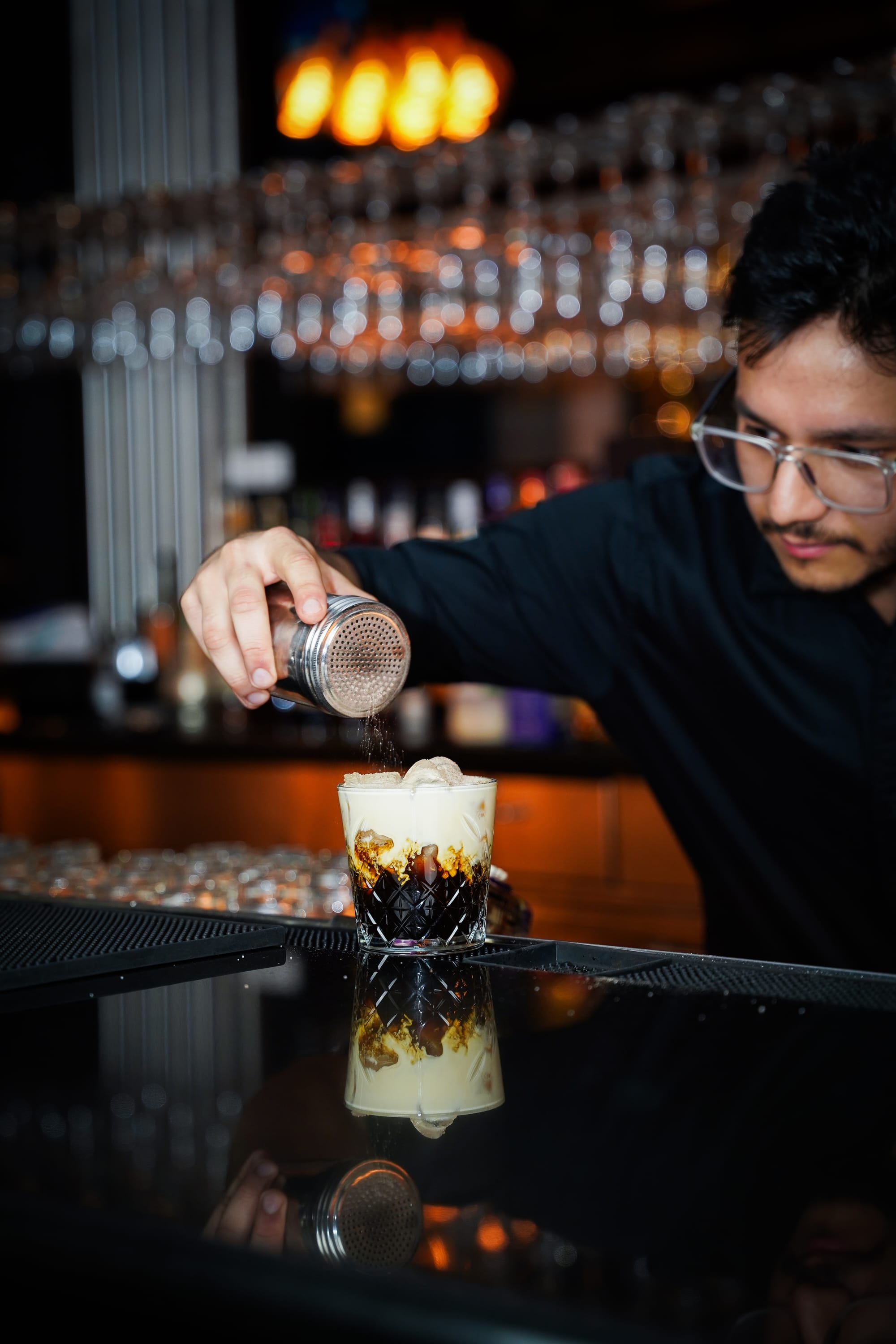 A bartender wearing glasses sprinkles spice over a creamy cocktail in a patterned glass at Soul bar, with the drink reflected on the glossy counter.