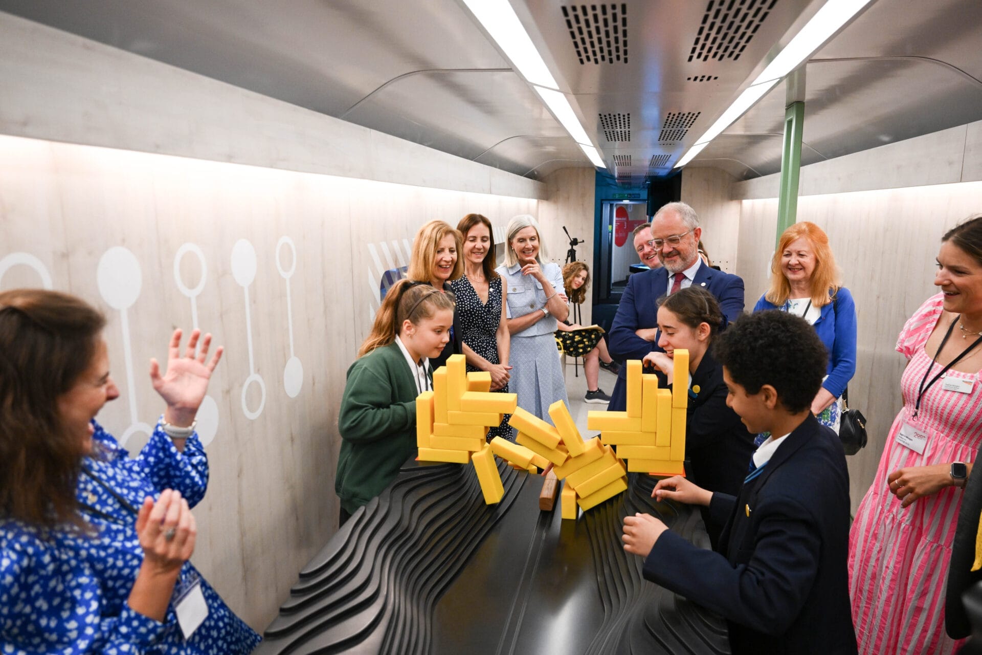 School children in uniform building structures with large yellow blocks inside the train carriage whilst adults observe and smile.