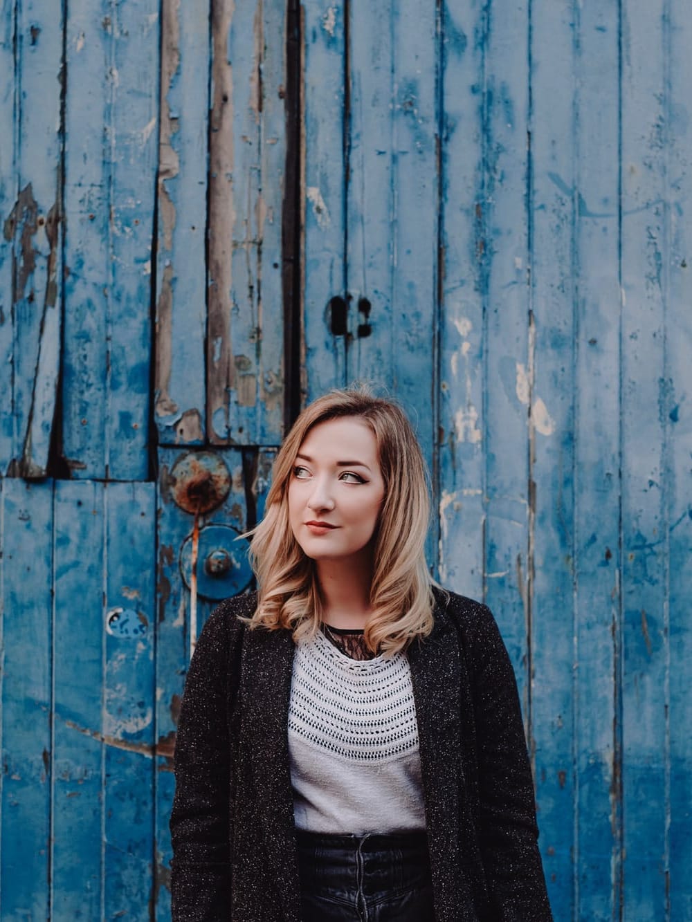 Iona Fyfe standing against a weathered blue painted wooden wall, wearing a dark sparkly cardigan over a white knit top.