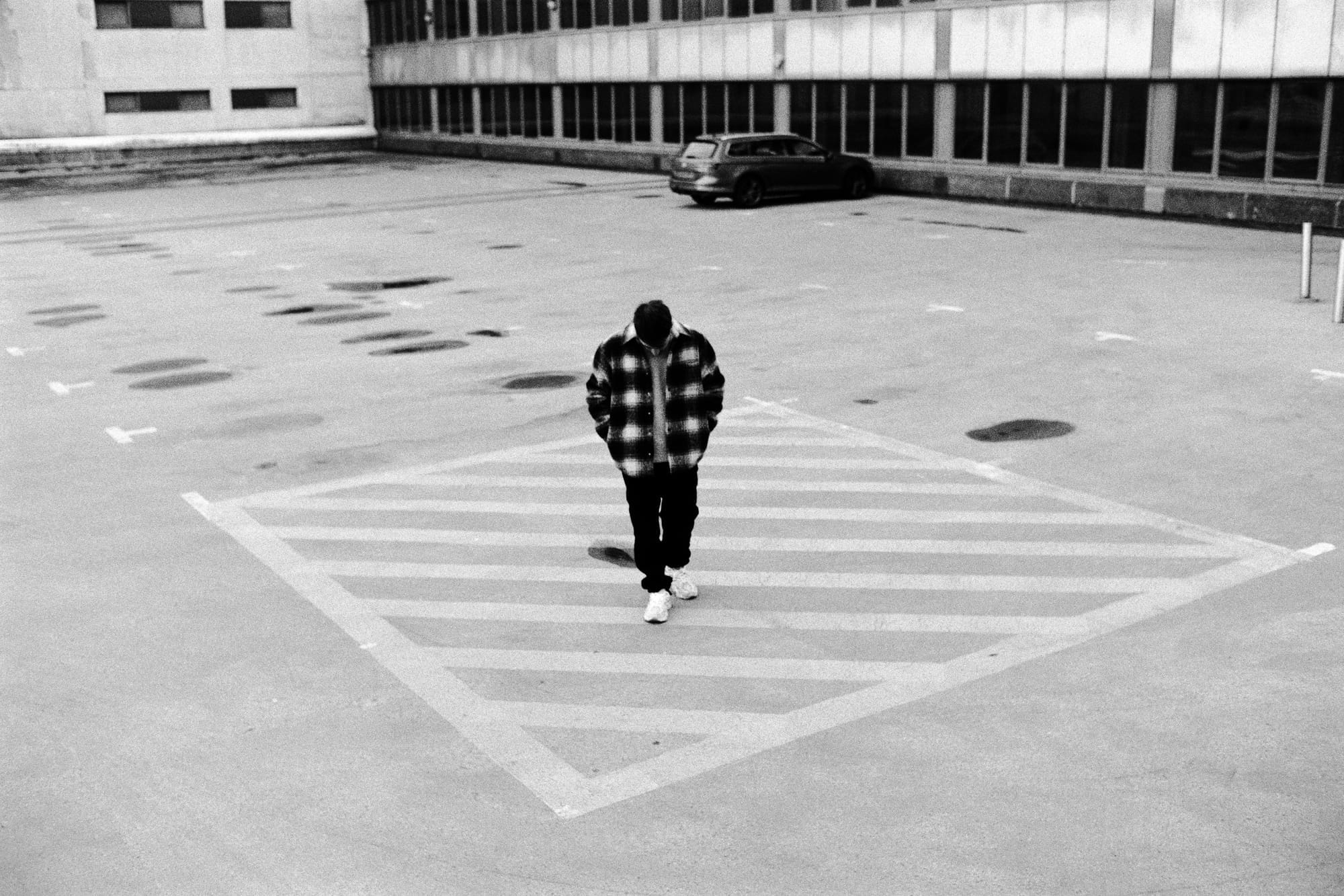 Solitary figure in checked jacket walking through empty car park with modernist buildings.