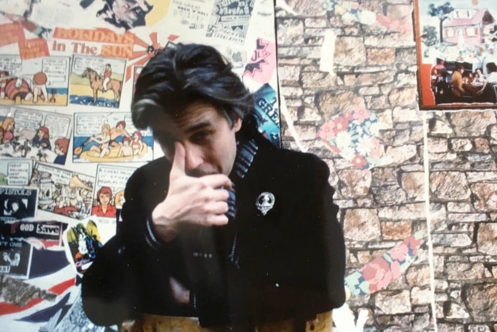 Jamie Reid in black clothing with hand raised to face, against wall covered with posters and artwork.