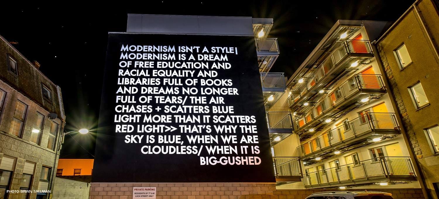 A large black wall mural with white capital letters displaying a poem about modernism, education, and the sky, installed on a building beside residential flats with lit windows at night.