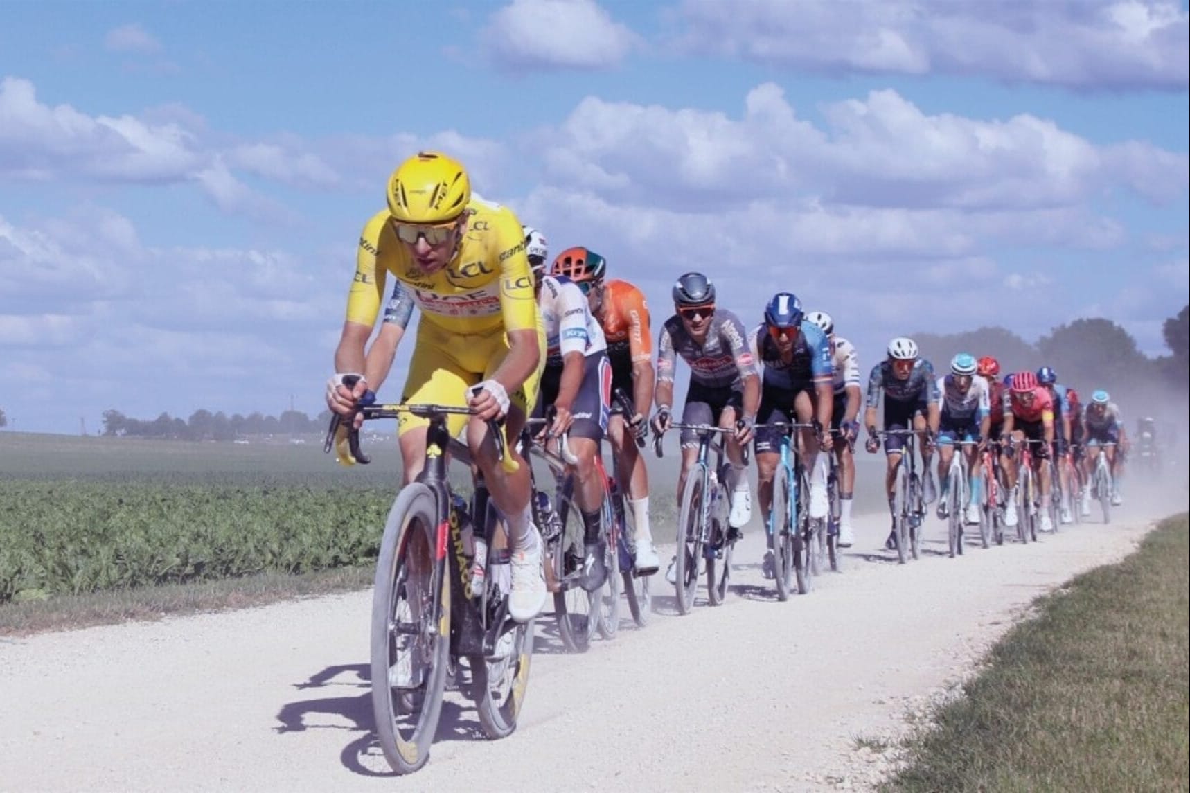 Peloton of cyclists led by rider in yellow jersey racing along dusty white road through green countryside under blue sky.