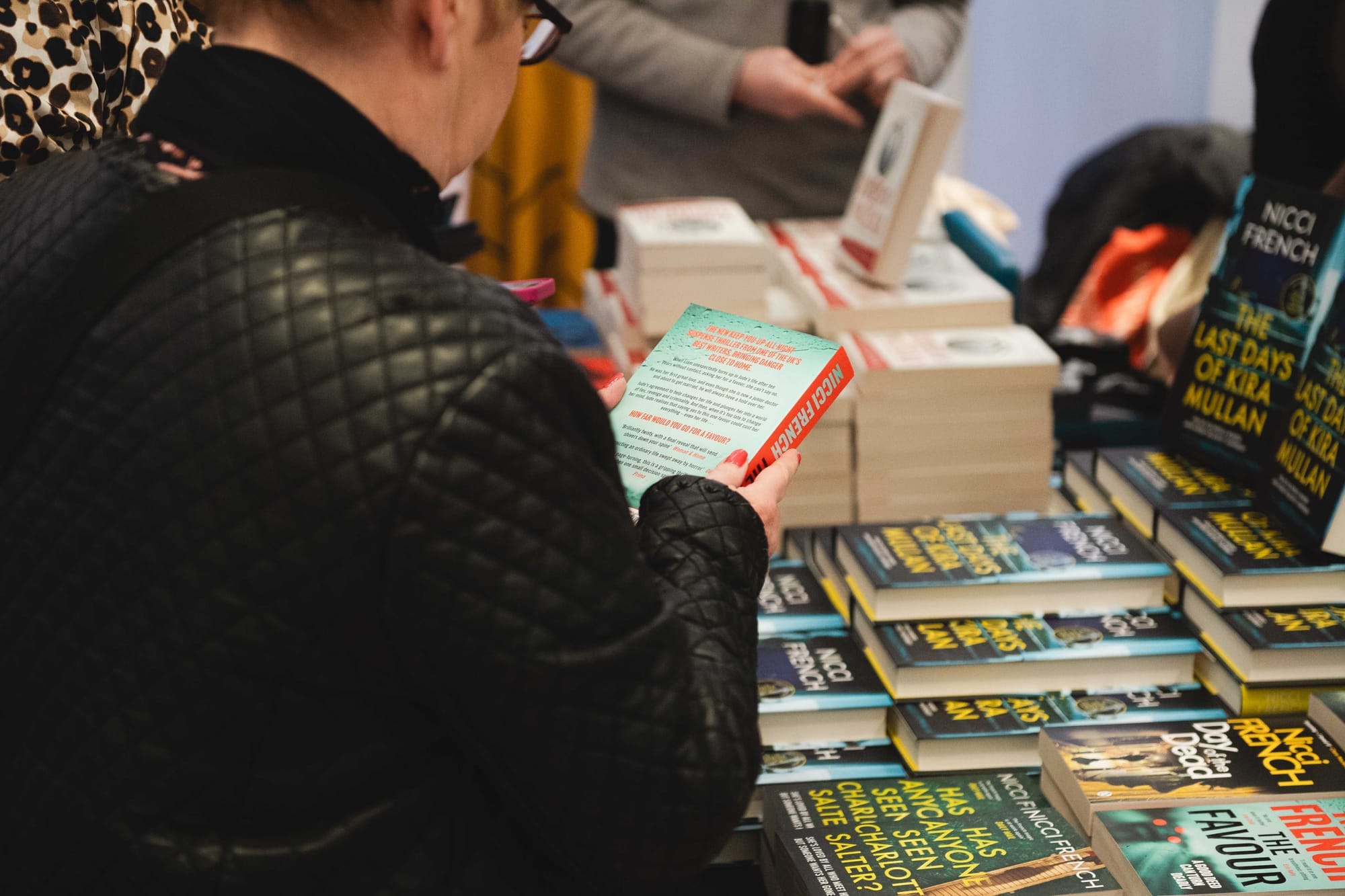 A reader in a black jacket examines a paperback at a table stacked with crime novels.