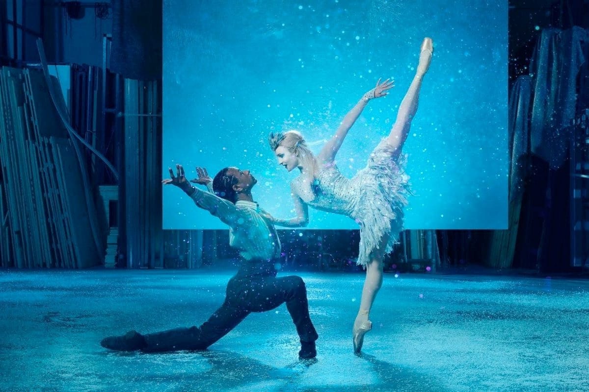 Ballet dancer in white feathered costume performing high kick beside kneeling partner in blue stage lighting.