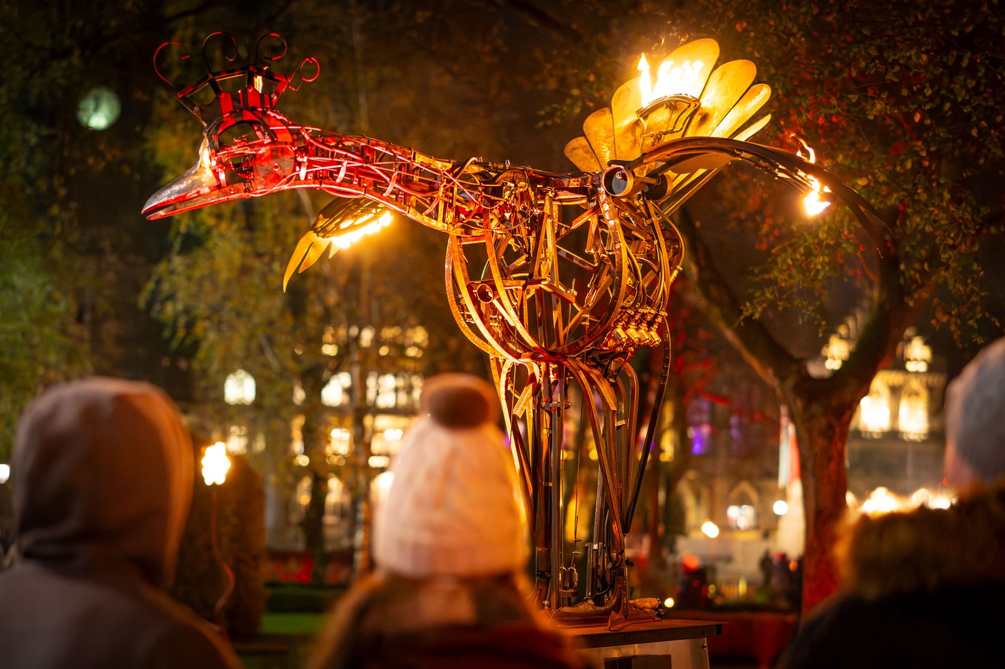Illuminated metal bird sculpture with golden wings and red head, breathing flames at night with crowd of onlookers in foreground and trees in background.