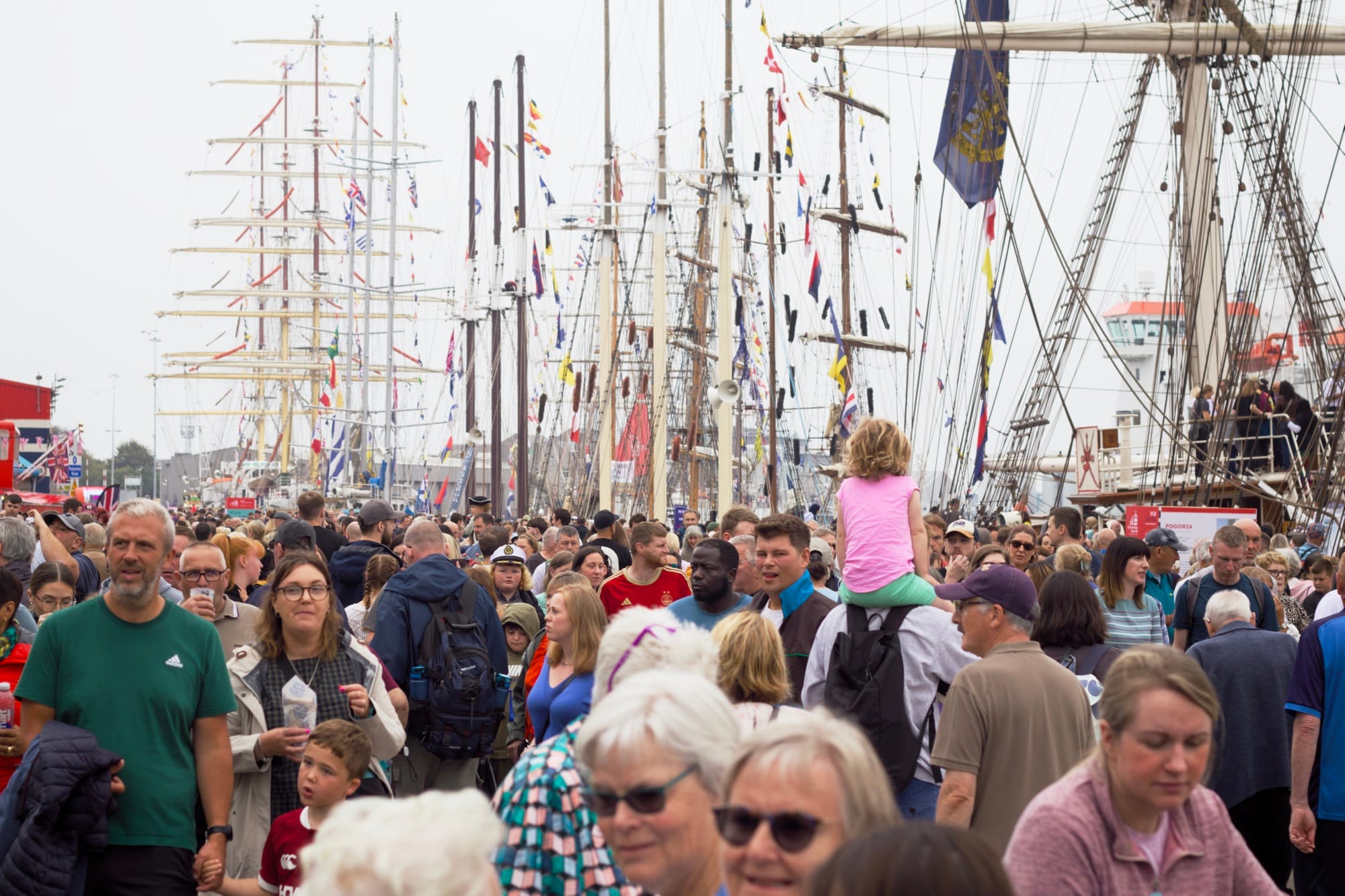 Crowds walking amongst tall ships with masts and rigging, child on adult's shoulders visible in pink top.