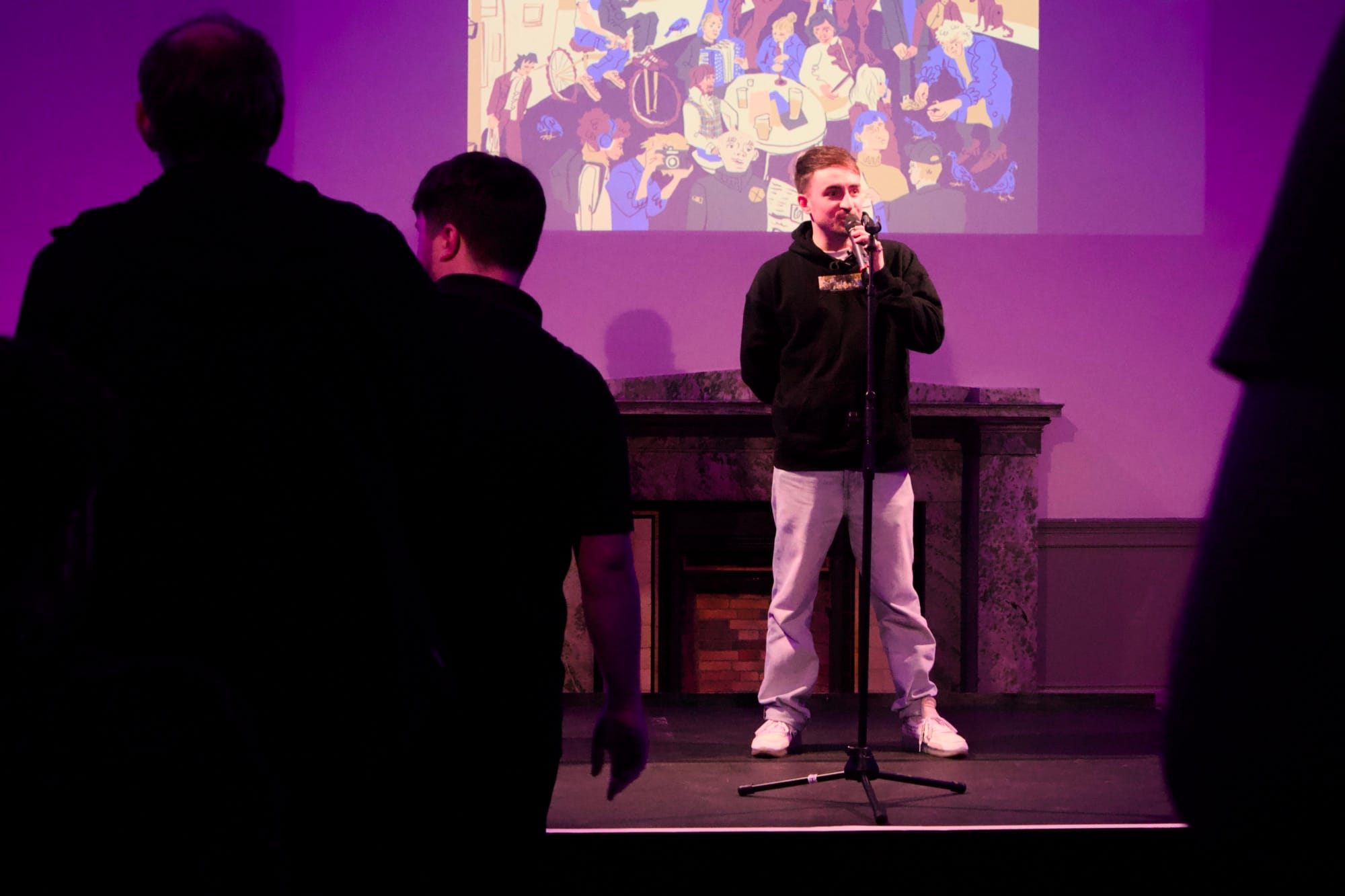 Vagrant Real Estste Person speaking into microphone on stage in purple-lit room with projected illustration behind.