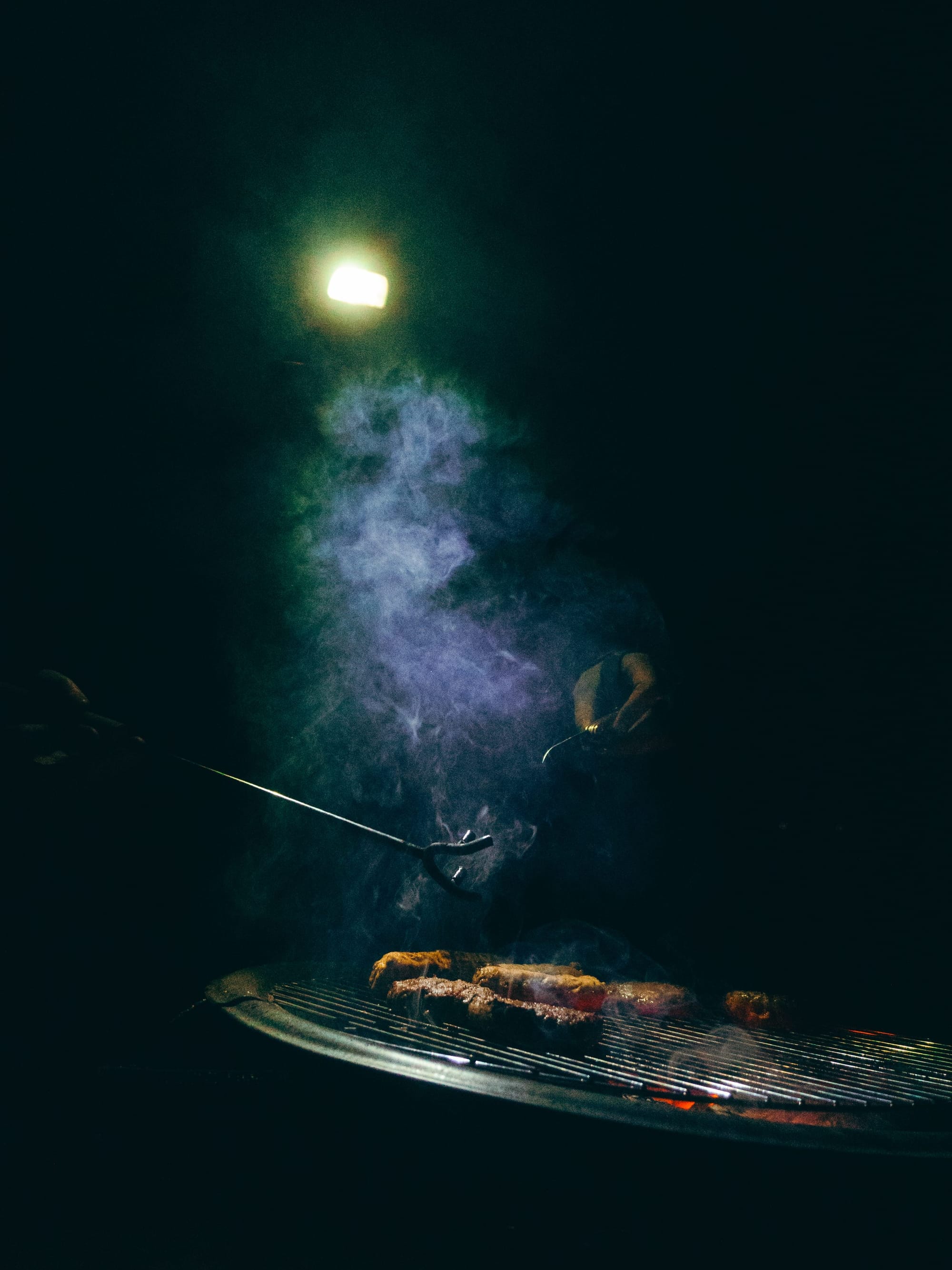 Sausages cooking on a barbecue at night, smoke rising into head torch glow