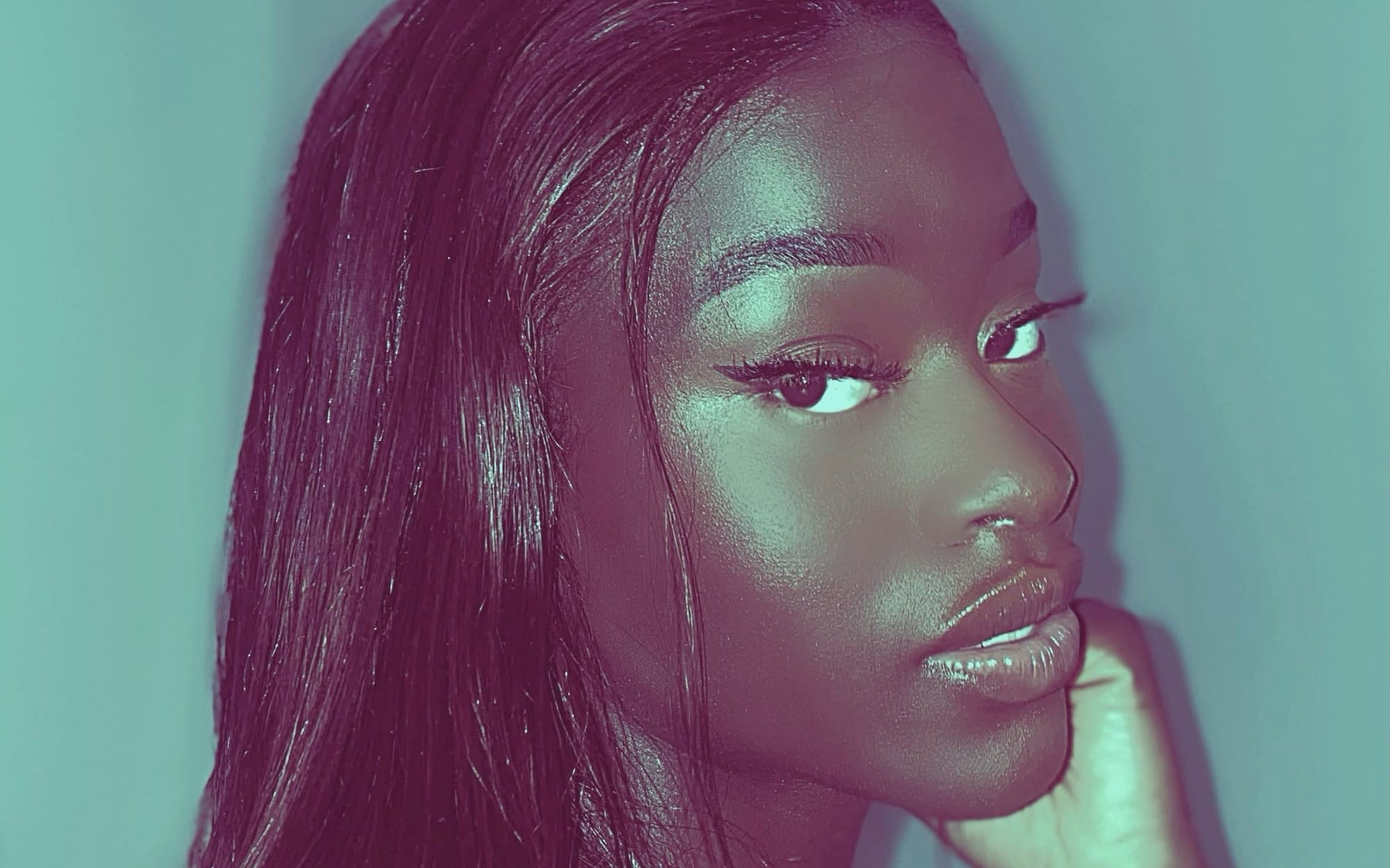 A close-up portrait of singer AiiTee against a teal background, hand raised to her chin.