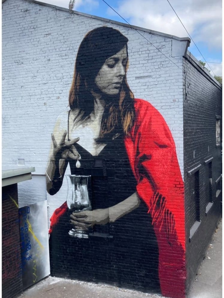 A large-scale stencil mural of a woman in a red shawl holding a silver vase.