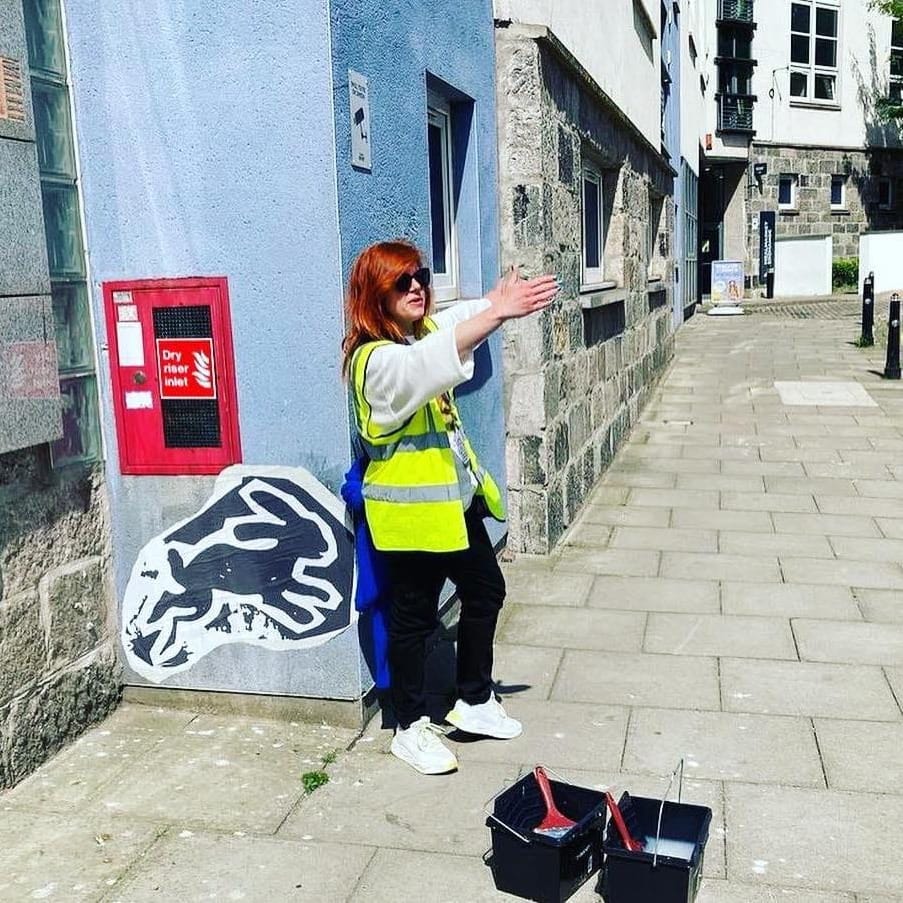 A woman in a hi-vis vest gestures at a black and white street art piece on an Aberdeen wall.