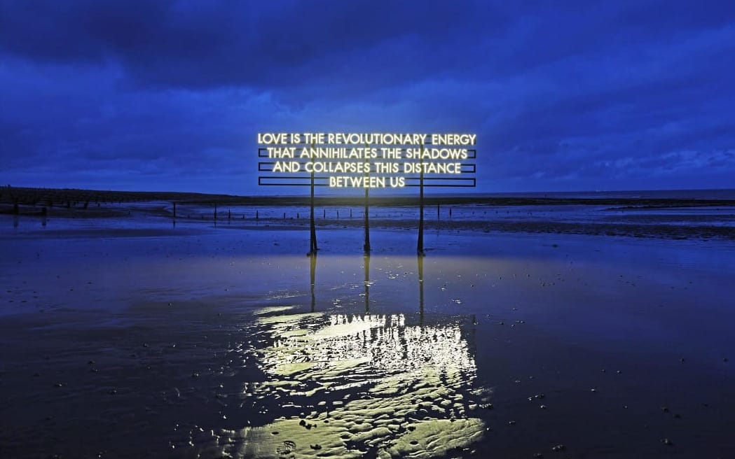 A illuminated text billboard on a beach at dusk, reflected in wet sand below.