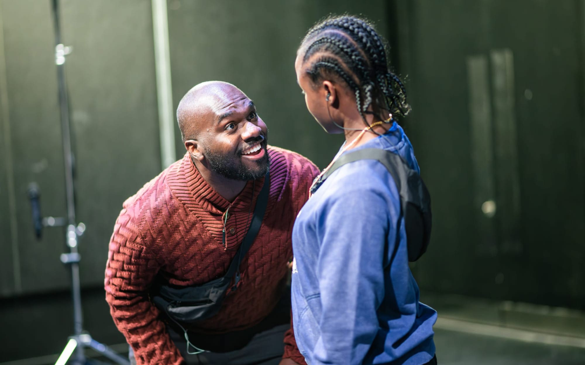Two actors face each other on a bare stage, one leaning in with a wide smile.