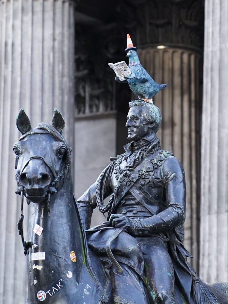 A bronze equestrian statue with a painted pigeon wearing a traffic cone on its head.
