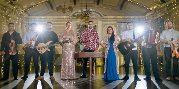 Eight musicians pose in a fairy-lit hall holding violins, guitar and accordion.