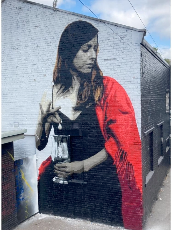 A large stencil mural of a woman in a red shawl holding a silver vase.