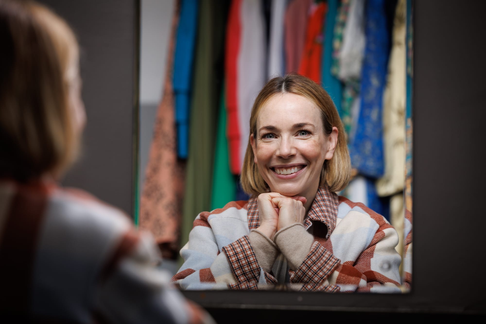 Laura Main smiling into a backstage dressing room mirror with costumes behind her
