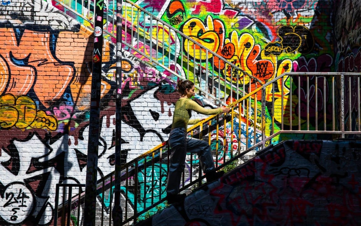 Alisa Oleva climbs a metal staircase against a graffiti-covered brick wall.