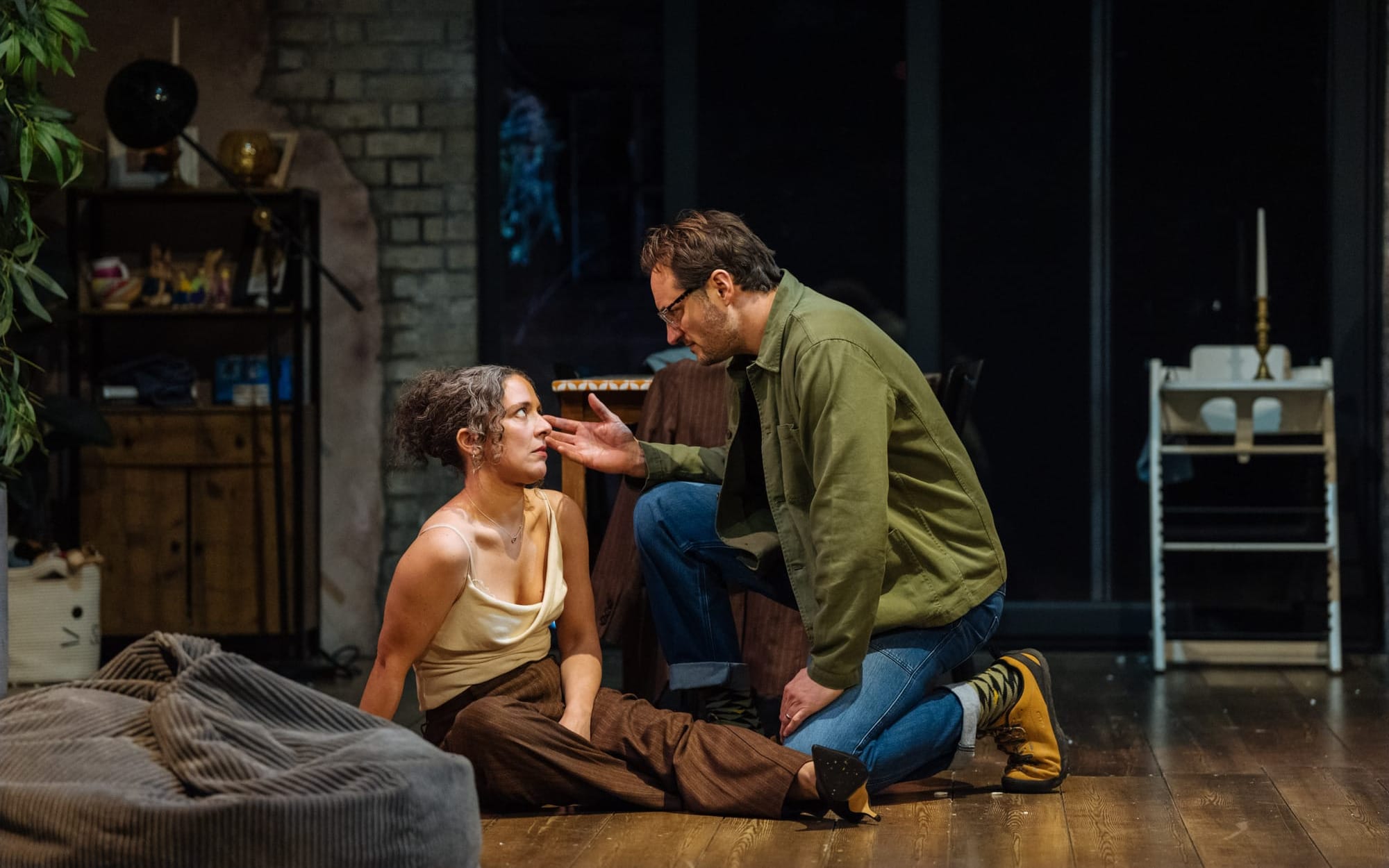 A woman sits on the floor as a man kneels beside her, touching her face.