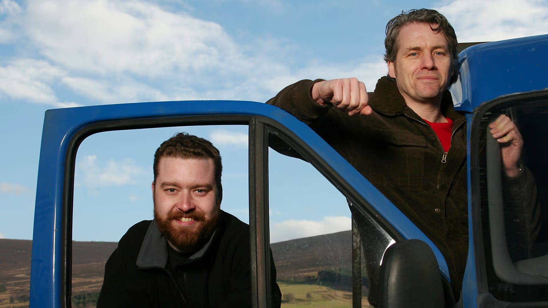 Two men pose in and around a blue truck against a Highland countryside backdrop.
