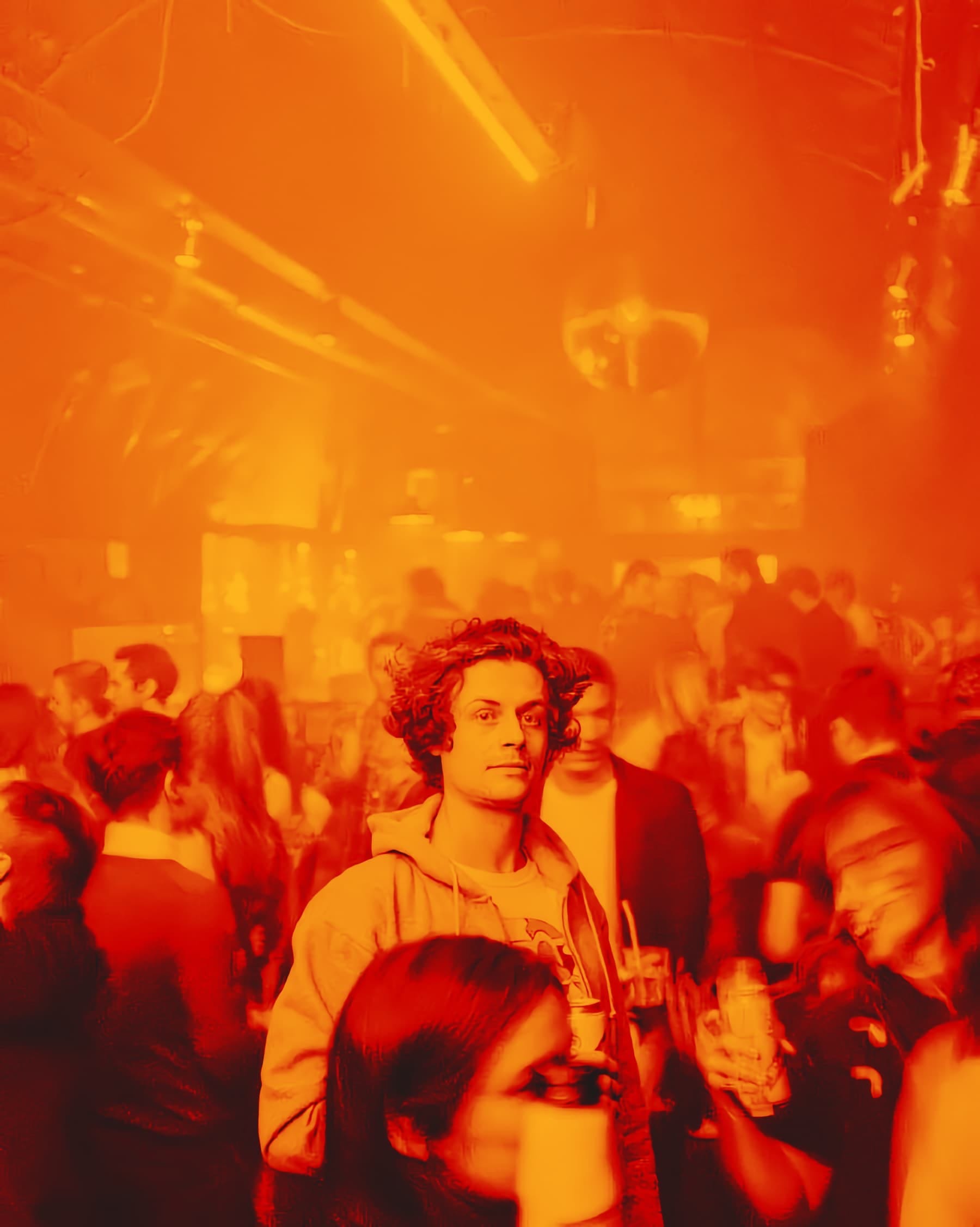 Orange-toned photo of a packed dance floor, a young man looking towards the camera. Caption: