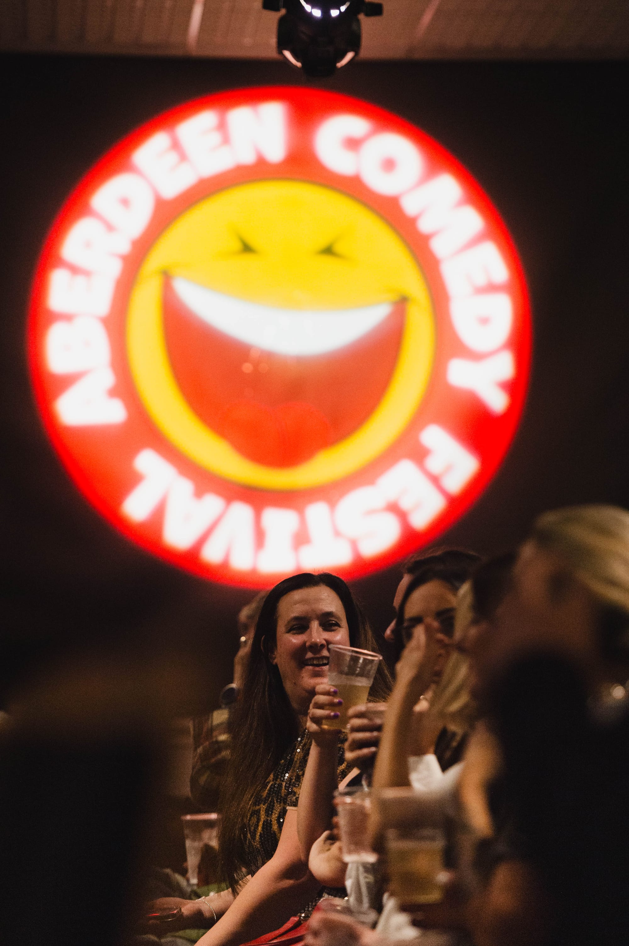 Crowd toasting drinks beneath the lit Aberdeen Comedy Festival smiley sign