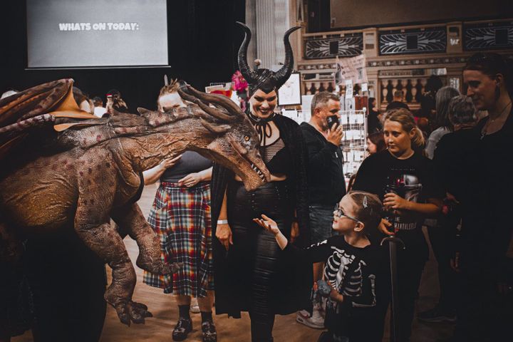 A child in a skeleton costume interacts with a dragon prop, surrounded by attendees including a Maleficent cosplayer.