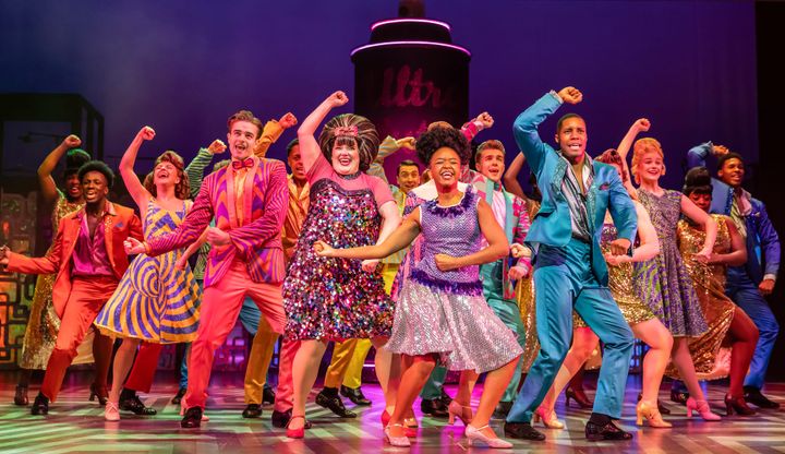 A colourful ensemble cast dances energetically in vibrant costumes on a brightly lit musical theatre stage.