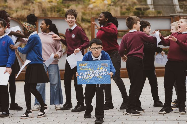 Child holding 'Playground Performance' sign, peers behind engaged in lively rehearsals.