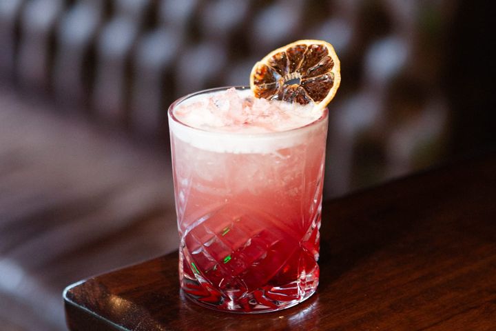 A pink cocktail in a cut-glass tumbler with ice and a dried citrus garnish on a wooden table.