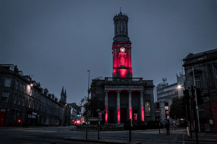 Aberdeen Arts Centre clock tower lit in red at dusk, surrounded by grey stone buildings and traffic.