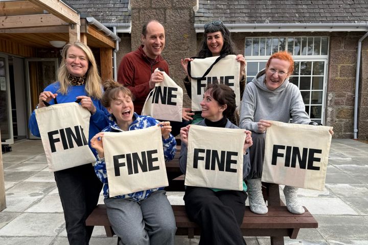 Group holding up canvas tote bags printed with the word “FINE” outside a building with stone walls.