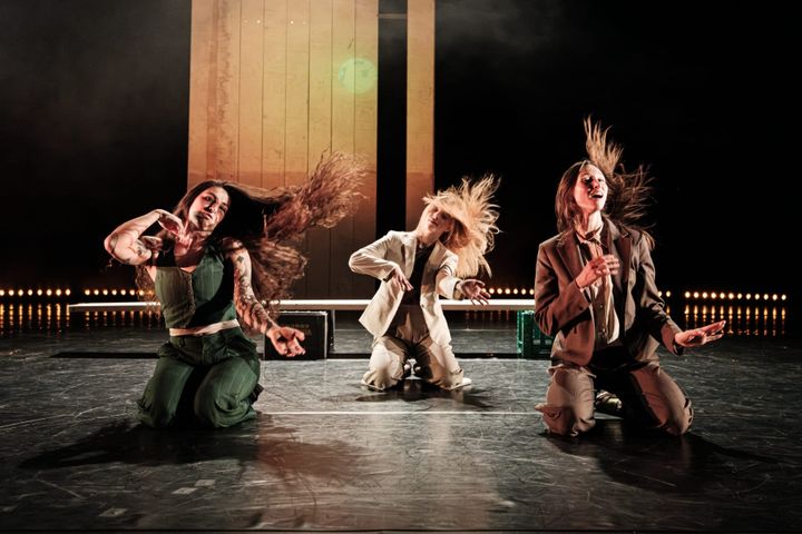 Three dancers on stage, mid-movement, with hair flying under dramatic lighting