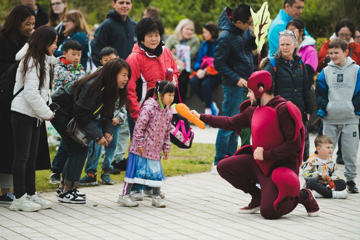 Performer in beetroot costume offers toy carrot to child, surrounded by smiling families outdoors.