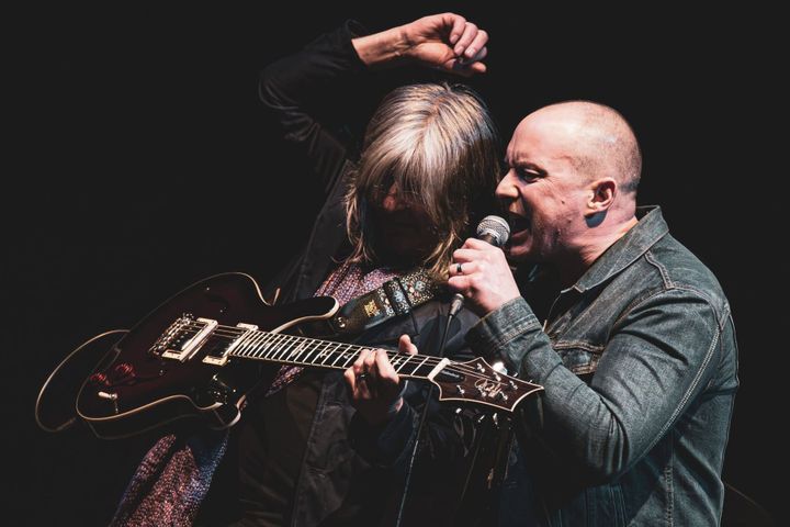 Gerry Jablonski and Nicky Aiken perform together, caught in a dramatic moment under dark stage lighting.