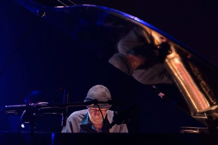 Musician Oscar Edelstein plays piano under stage lights, reflected in the instrument’s lid.