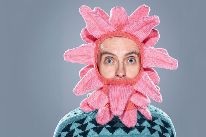 Greg Sinclair wears a bright pink knitted headpiece shaped like petals over a patterned jumper.