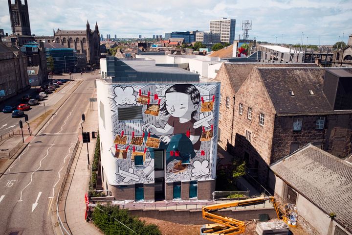 A mural beside an Aberdeen road of a cartoon character pinning photos to a washing line against a city background.