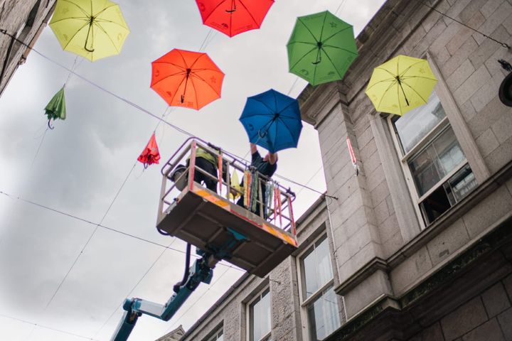 Two workers on a lift install colourful umbrellas overhead between granite buildings in Aberdeen city centre.