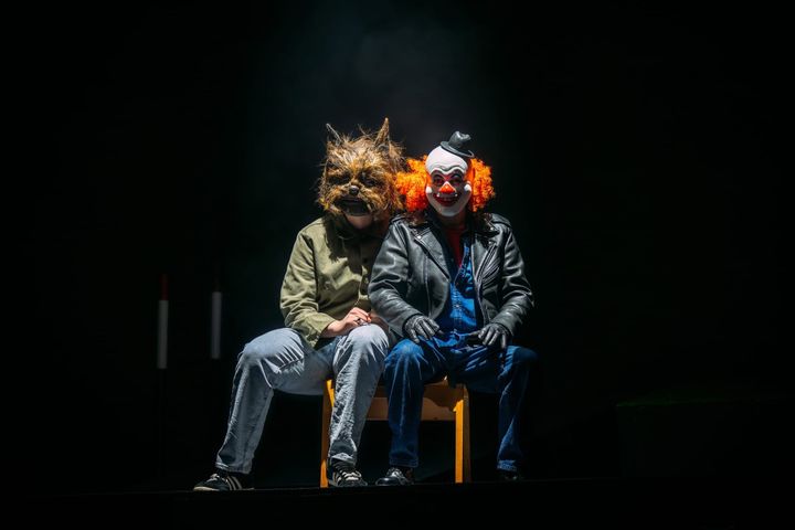 Two people sit in darkness wearing a werewolf and clown mask, lit by a dramatic spotlight.